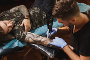 professional tattooer artist doing tattoo arm young man by machine with black ink
