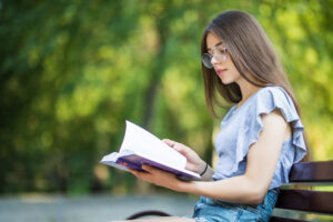 side view pleased brunette woman eyeglasses sitting bench reading book park