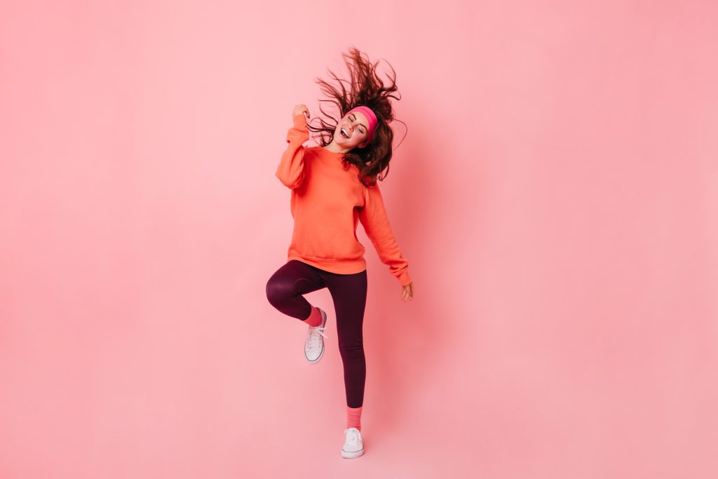 Lady in bright sweatshirt and brown leggings dances against pink background. Fitness trainer enjoying workout.
