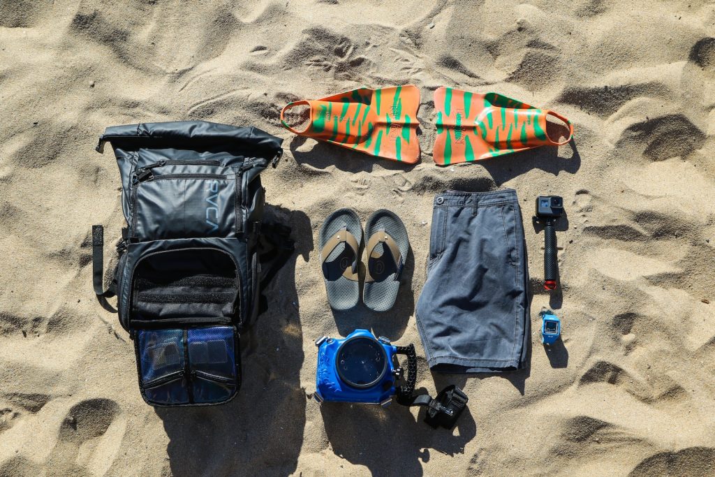 Sand Bag flat-lay photography of flip-flops, hiking backpack, board, shorts, and action camera